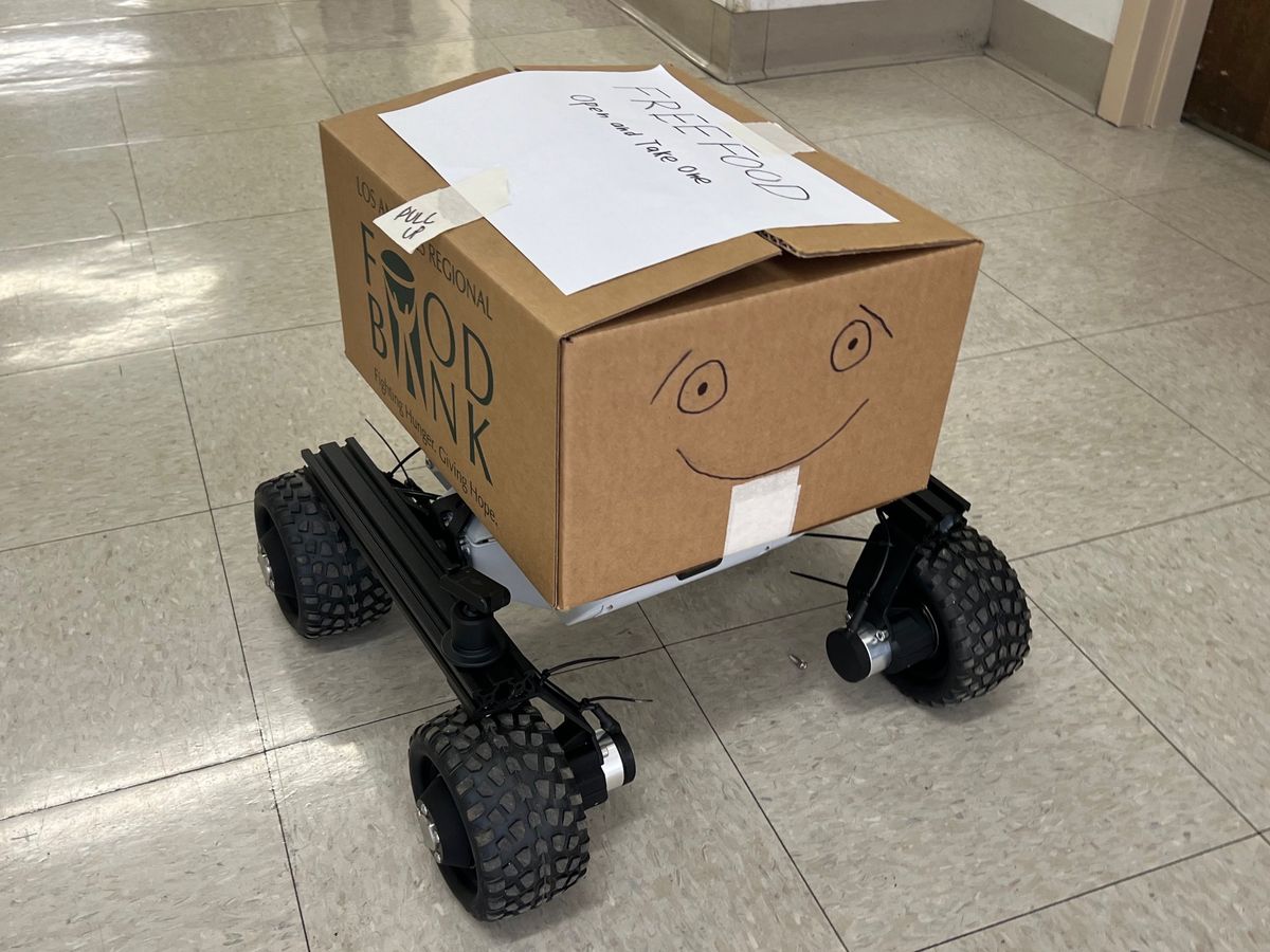 Food Angel: a cardboard box with a drawn smiley face and a handwritten FREE FOOD sign, on a wheeled robot base