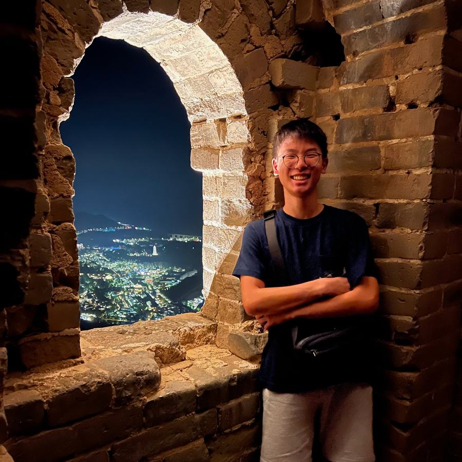 Ethan leaning against the stone wall of a Great Wall watchtower at night, with city lights through the arched window