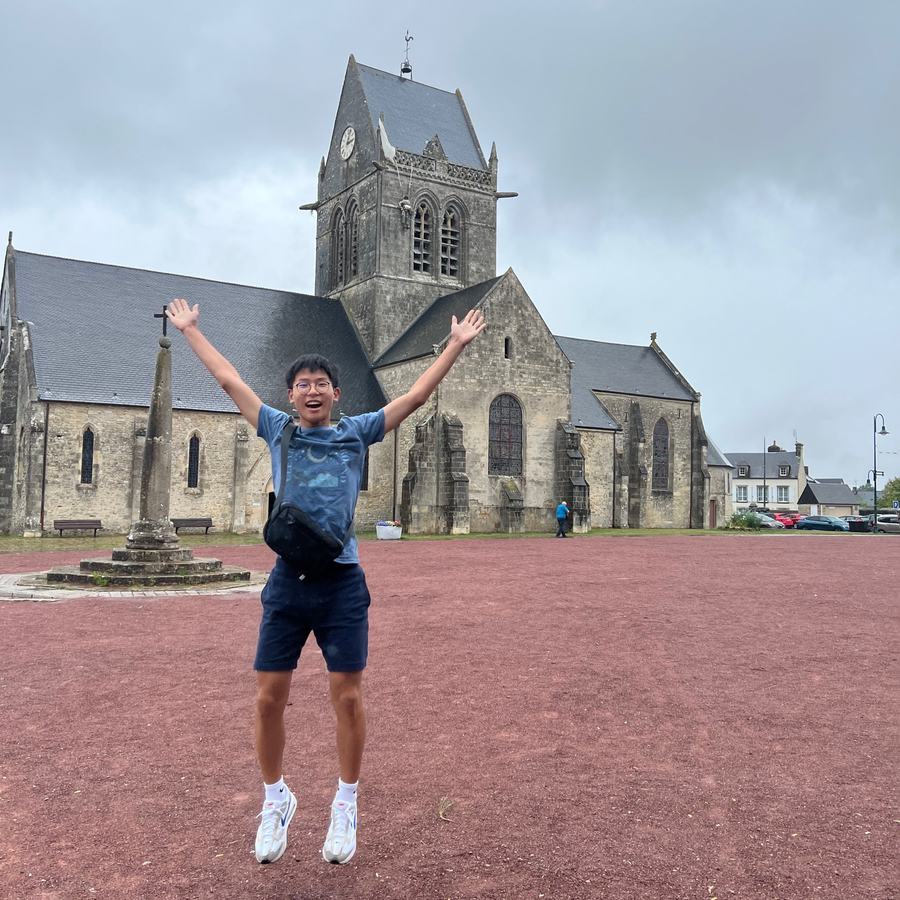 Ethan jumping with his arms raised in front of the church of Sainte-Mère-Église, with a paratrooper figure hanging from the steeple