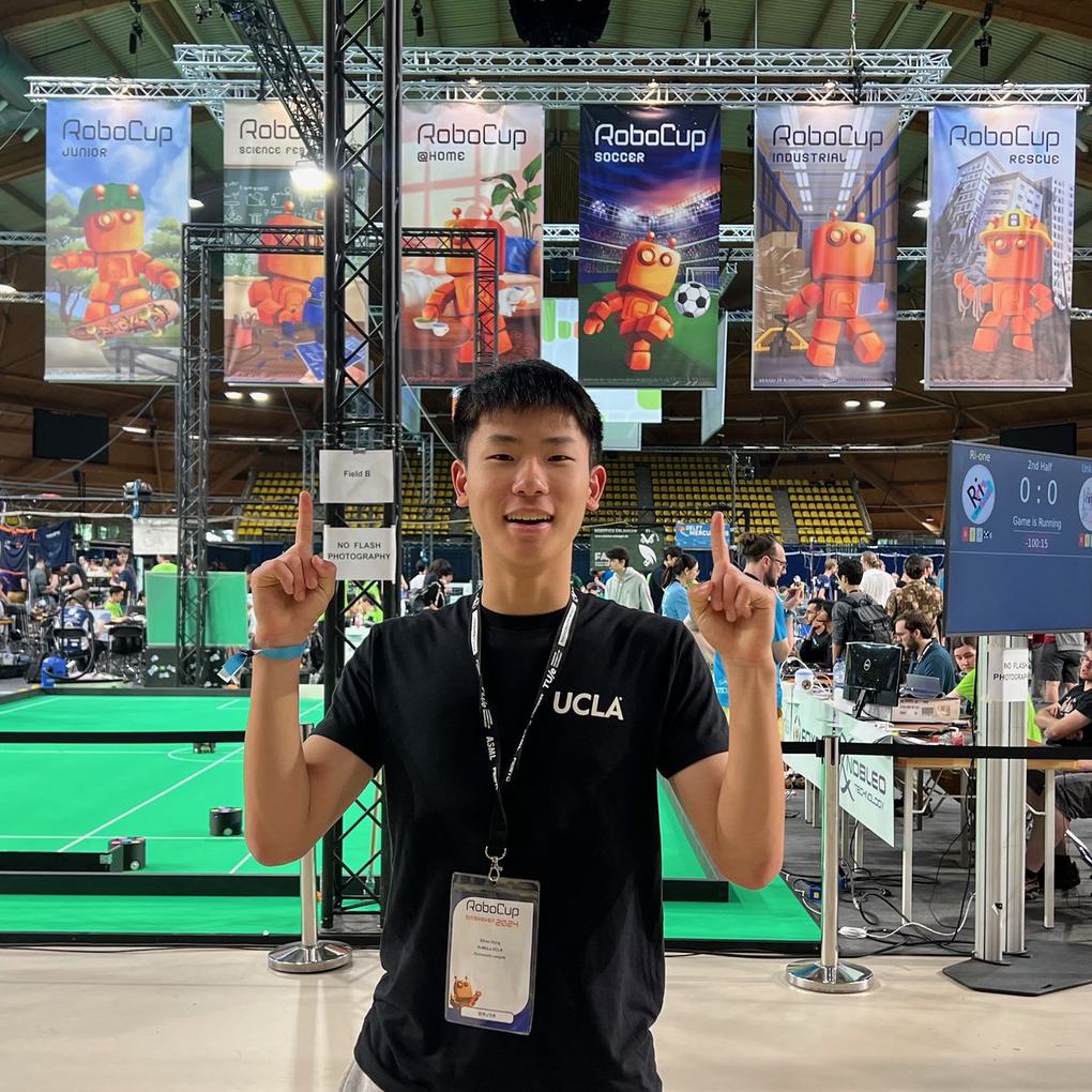 Ethan pointing up with both index fingers under RoboCup banners in the Eindhoven competition hall