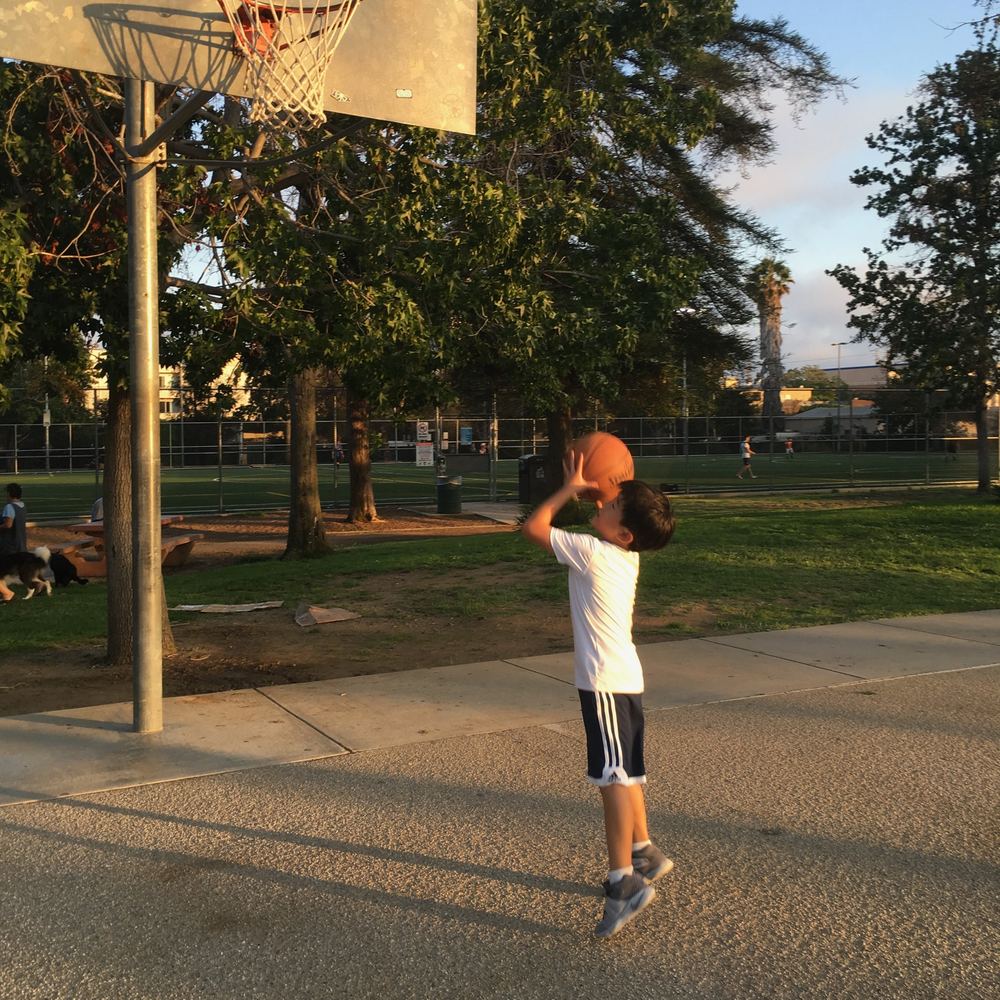 Young Ethan in a white shirt shooting a basketball at an outdoor park hoop