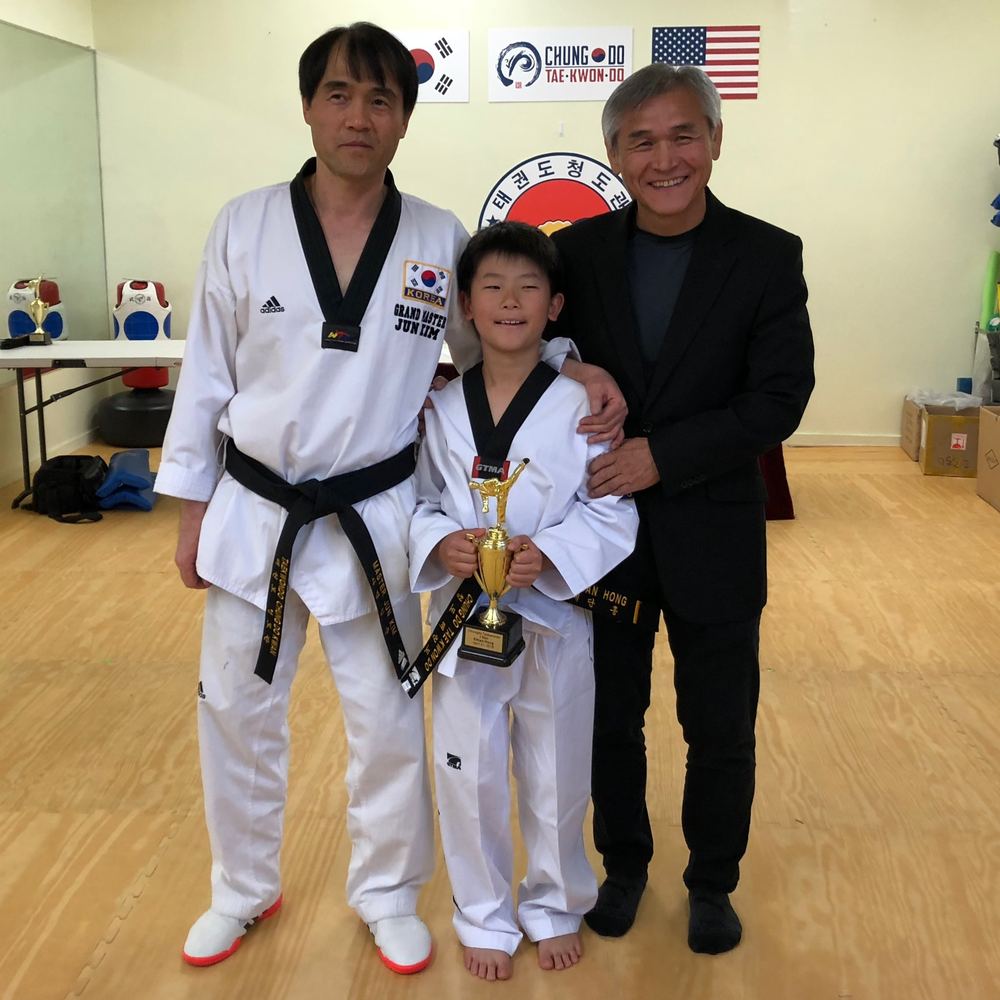 Ethan in his new black belt holding a trophy, standing with Grand Master Jun Kim