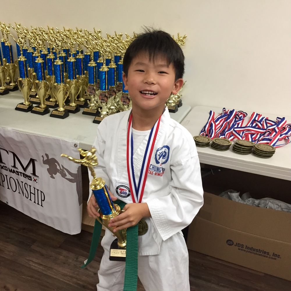 Young Ethan smiling in his uniform and green belt, holding a tournament trophy with a medal around his neck